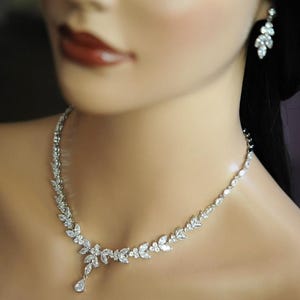 May include: A silver necklace and matching earrings set, featuring a leaf design with clear crystals. The necklace has a pendant drop. The earrings are a similar leaf design. The jewellery is displayed on a mannequin.