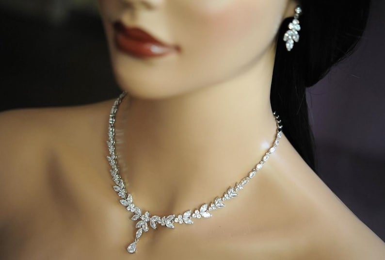 May include: A silver necklace and matching earrings set, featuring a leaf design with clear crystals. The necklace has a pendant drop. The earrings are a similar leaf design. The jewellery is displayed on a mannequin.