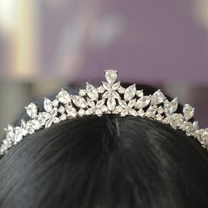 May include: A silver tiara adorned with clear, sparkling crystals. The tiara showcases a floral design with diverse crystal shapes, including teardrops and marquise cuts. The tiara is set against a dark backdrop, accentuating its sparkle. Ideal for weddings or special events.