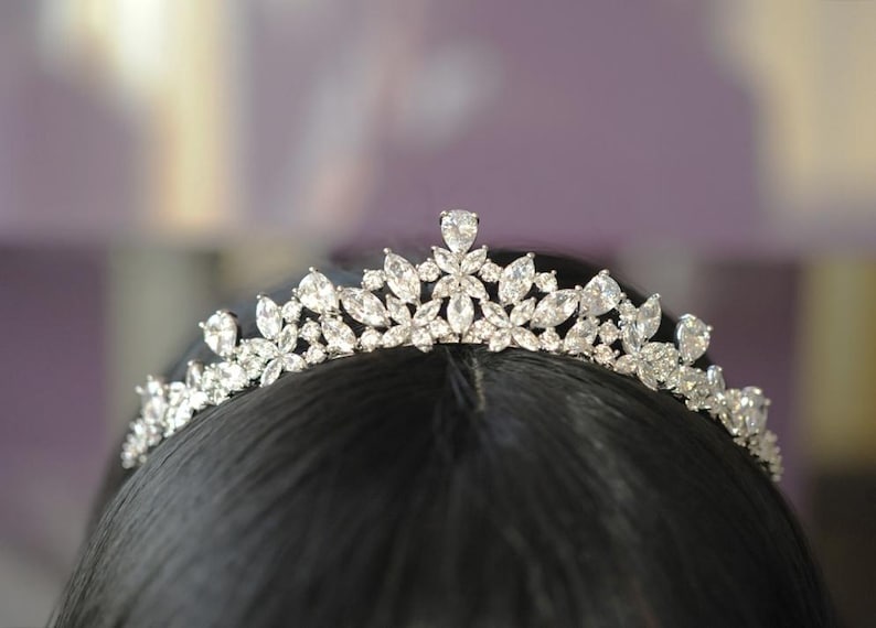 May include: A silver tiara adorned with clear, sparkling crystals. The tiara showcases a floral design with diverse crystal shapes, including teardrops and marquise cuts. The tiara is set against a dark backdrop, accentuating its sparkle. Ideal for weddings or special events.
