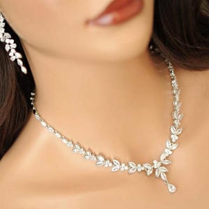 May include: A silver necklace and matching earrings set, adorned with sparkling clear crystals. The necklace has a floral design with a teardrop pendant. The earrings are a cascading floral design. The jewellery is worn on a person's neck and ear.