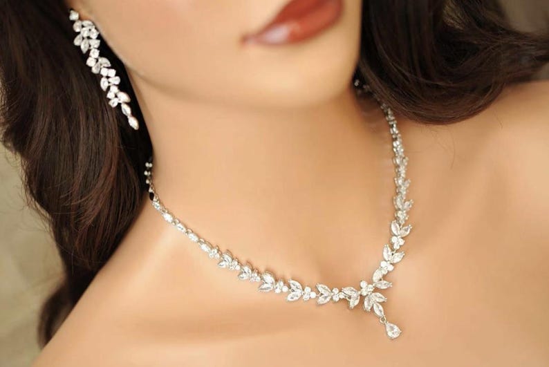 May include: A silver necklace and matching earrings set, adorned with sparkling clear crystals. The necklace has a floral design with a teardrop pendant. The earrings are a cascading floral design. The jewellery is worn on a person's neck and ear.