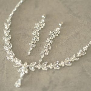 May include: A silver necklace and earring set with a leaf design. The jewellery features clear, brilliant-cut stones. The necklace has a teardrop pendant. The earrings are long and dangle, creating a glamorous look.