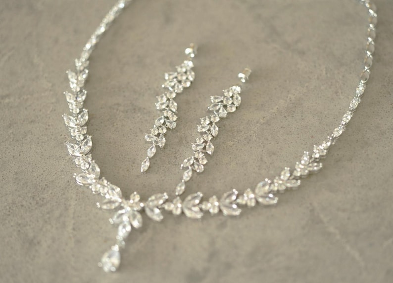 May include: A silver necklace and earring set with a leaf design. The jewellery features clear, brilliant-cut stones. The necklace has a teardrop pendant. The earrings are long and dangle, creating a glamorous look.