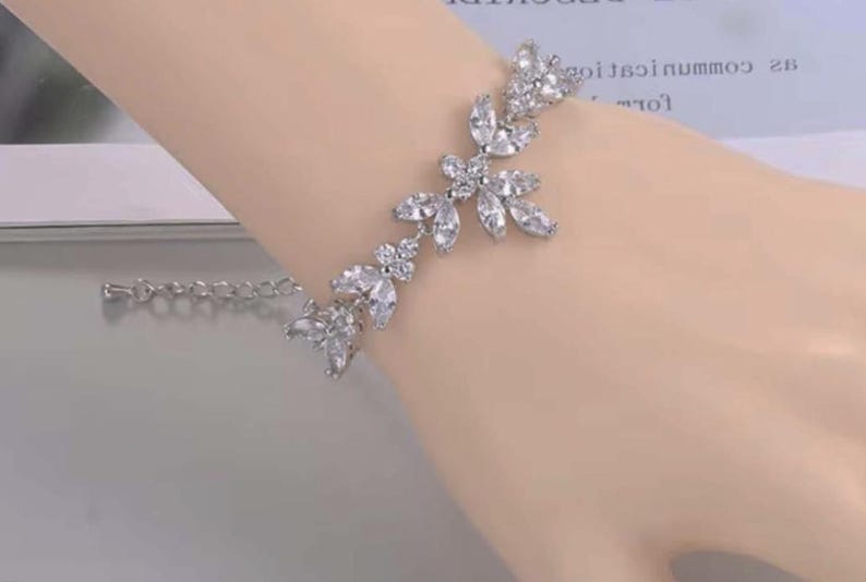 May include: Silver bracelet with a floral design, adorned with clear, sparkling crystals. The bracelet includes a delicate chain for adjustable sizing. The crystals are arranged in a leaf-like pattern, creating an elegant accessory.
