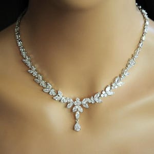 May include: A silver necklace with a floral design, adorned with clear crystals. The necklace features a leaf-like pattern of round and marquise-cut crystals. A teardrop-shaped crystal pendant hangs from the centre. The necklace is worn around the neck.