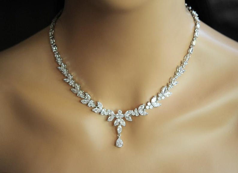 May include: A silver necklace with a floral design, adorned with clear crystals. The necklace features a leaf-like pattern of round and marquise-cut crystals. A teardrop-shaped crystal pendant hangs from the centre. The necklace is worn around the neck.