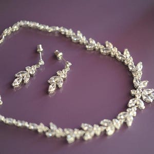 May include: A silver necklace and earring set with clear crystal embellishments. The necklace and earrings feature a floral and leaf design. The jewellery is presented on a purple surface, showcasing the sparkling crystals.