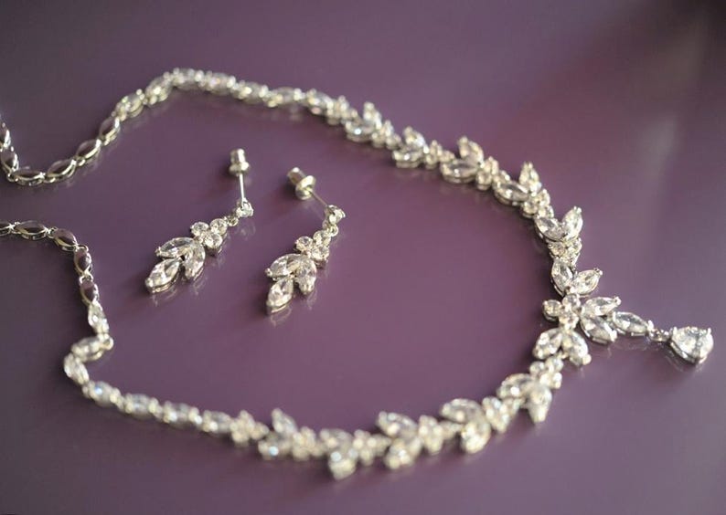 May include: A silver necklace and earring set with clear crystal embellishments. The necklace and earrings feature a floral and leaf design. The jewellery is presented on a purple surface, showcasing the sparkling crystals.
