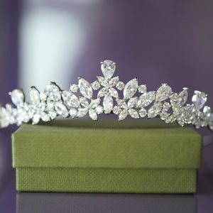 May include: A silver tiara featuring a floral pattern, embellished with many clear, sparkling crystals. The tiara rests on a small, green, textured box, set against a blurred purple backdrop.