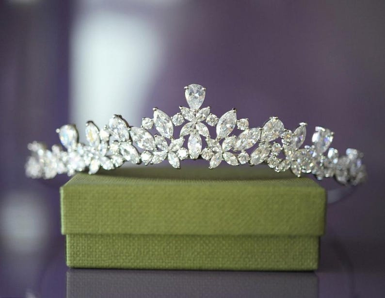 May include: A silver tiara featuring a floral pattern, embellished with many clear, sparkling crystals. The tiara rests on a small, green, textured box, set against a blurred purple backdrop.