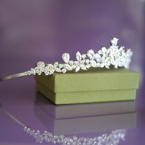 May include: A silver tiara with clear gemstones in a floral pattern. It rests on an olive-green box, reflecting on a purple surface. The gemstones are of different sizes and shapes, creating a sparkling effect.