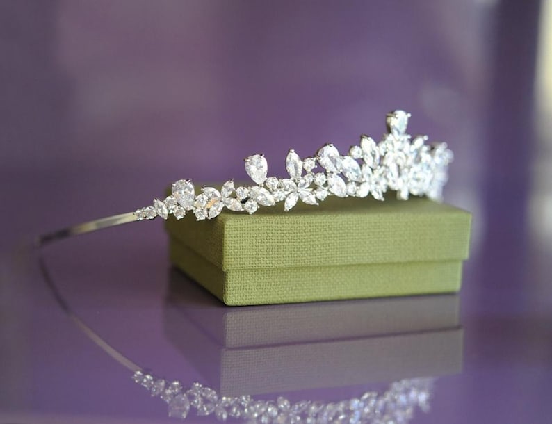 May include: A silver tiara with clear gemstones in a floral pattern. It rests on an olive-green box, reflecting on a purple surface. The gemstones are of different sizes and shapes, creating a sparkling effect.