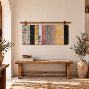 Könnte beinhalten: Ein rechteckiger Wandbehang mit vertikalen Streifen in verschiedenen Farben und Mustern, der an einer Holzstange mit Lederriemen befestigt ist. Das Textil zeigt Abschnitte in Schwarz, Gelb, Rot, Grau und Blau mit einer abgenutzten Textur.