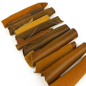 May include: An assortment of leather hides in various shades of brown, from light tan to deep chocolate. Some are rolled, while others are folded, showcasing the texture and color variations. The hides are arranged diagonally against a white background.