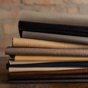 May include: A stack of various textured leather swatches in shades of gray, black, tan, and brown. The leather samples are neatly arranged, showcasing the different colors and textures available for upholstery or crafting projects.