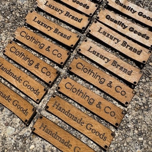 May include: A collection of rectangular, brown leather tags with embossed text. The tags display phrases like "Luxury Brand," "Clothing & Co.," and "Handmade Goods." The tags have small holes at each end.