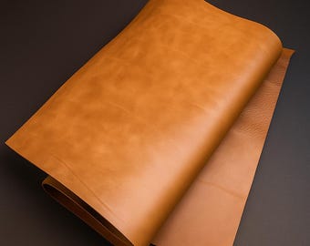 May include: A rolled sheet of tan-colored leather, with a smooth, slightly glossy surface. The leather is folded over, revealing a second sheet of the same material. The background is a dark gray.