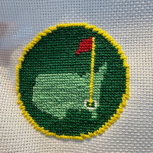 Op de afbeelding: Een handgemaakt kruissteek golfbaan embleem. Het ontwerp toont een groene golfbaan kaart met een rode vlag op een gele paal. Het embleem heeft een gele rand en is geborduurd op witte stof.