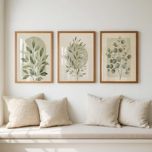 May include: Three framed wall art prints featuring watercolor botanical illustrations of green leaves and eucalyptus branches on a cream background, each enclosed in a light brown wooden frame, displayed in a horizontal row above four beige pillows.