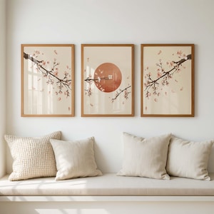 May include: Three framed art prints featuring a cherry blossom branch with pink flowers and falling petals. The middle print has a large orange sun. The frames are light brown, and the background is a cream color.