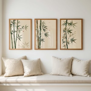May include: Three framed bamboo art prints. Each print features a watercolour illustration of bamboo stalks and leaves in shades of green and brown against a beige background. The frames are made of light brown wood. The prints are displayed above a row of beige cushions.