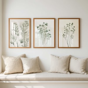 以下が含まれることがあります： 緑色の植物と花の水彩イラストが描かれた、額装された3つのボタニカルプリント。プリントは薄茶色の木製フレームに入っています。ベージュのクッションが、アートワークの下のベンチに置かれています。