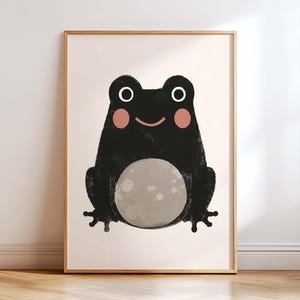 May include: A framed art print featuring a whimsical illustration of a black frog with a light gray belly and pink cheeks. The frog has white eyes and a smiling mouth. The artwork is set against a light beige background.