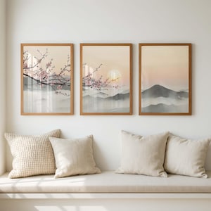 Puede incluir: Tres impresiones enmarcadas de un paisaje montañoso con flores de cerezo. La obra de arte utiliza una paleta suave de beige, rosa y gris. Los marcos son de color marrón cálido. Las impresiones se exhiben sobre un banco con cojines.