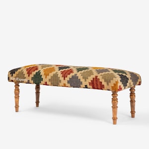 Puede incluir: Un banco rectangular con una superficie de tela tejida que presenta un patrón geométrico de diamantes multicolor en tonos rojo, amarillo, verde y gris. El banco se apoya sobre cuatro patas de madera torneada con un acabado marrón pulido.