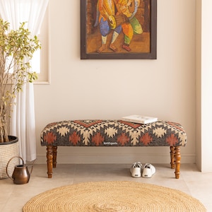 May include: Upholstered bench with a geometric pattern in shades of brown, beige, and red. The bench has turned wooden legs and a rectangular shape. A book and a pair of white trainers are on the bench.