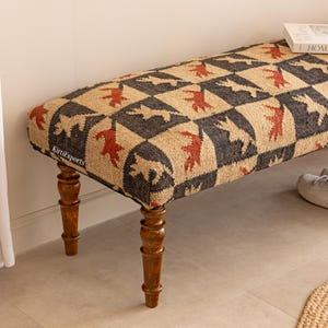 Puede incluir: Un banco rectangular con cuatro patas de madera torneada en acabado marrón oscuro. El asiento está tapizado en una tela tejida con un patrón de cuadros negros y beige, cada uno con una figura animal naranja estilizada. El texto dice KirtiExports.