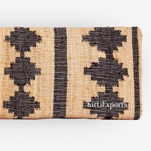 Puede incluir: Un tejido rectangular con un patr&oacute;n repetitivo de formas geom&eacute;tricas de diamante en negro y rayas verticales sobre un fondo beige natural. La tela tiene una textura gruesa visible e incluye el texto en blanco KirtiExports.
