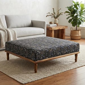 Pode incluir: Um otomano quadrado com estofamento de tecido escuro com um padrão floral repetitivo em tons suaves de azul, dourado e creme. A peça é sustentada por quatro pernas de madeira torneada com acabamento em castanho médio.