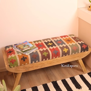 Puede incluir: Un banco de madera con un asiento rectangular tapizado en tela tejida con un patrón geométrico multicolor de cuadrados y cruces en rojo, verde, amarillo y azul. Una pila de libros descansa sobre el asiento y el texto KirtiExports es visible.