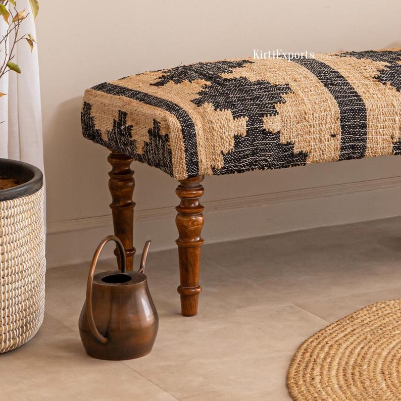 Puede incluir: Un banco rectangular con un asiento de yute tejido con un patr&oacute;n geom&eacute;trico en negro y beige. El banco se apoya sobre cuatro patas de madera torneada de color marr&oacute;n oscuro. El texto KirtiExports es visible en la parte superior derecha.