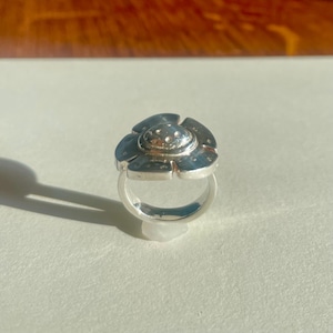 以下が含まれることがあります： 花型のデザインのシルバーリング。リングは、小さな均等に配置されたドットが付いた、中央の隆起した円形要素と、6つの花びらのような形状で構成されています。リングはシンプルなバンドにセットされています。