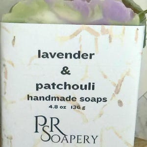 May include: A handmade soap bar with a white label featuring the text "lavender & patchouli handmade soaps". The soap is 4.8 oz (136 g) and has a marbled purple and green top. The soap is displayed on a wooden block.