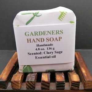 May include: A bar of handmade "Gardeners Hand Soap" on a wooden soap dish. The soap is wrapped in white paper with green gardening tool illustrations. The label indicates it is scented with clary sage essential oil and weighs 4.8 oz.