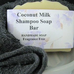 May include: A light-colored Coconut Milk Shampoo Soap Bar, wrapped in a label with floral accents, rests on a white ceramic soap dish. The bar is nestled in a dark gray, textured towel. The label reads "HANDMADE SOAP Fragrance Free."