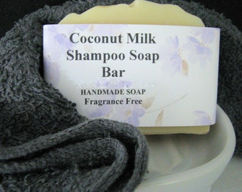 May include: A light-colored Coconut Milk Shampoo Soap Bar, wrapped in a label with floral accents, rests on a white ceramic soap dish. The bar is nestled in a dark gray, textured towel. The label reads "HANDMADE SOAP Fragrance Free."