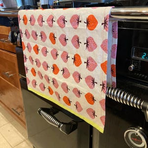 Könnte beinhalten: Ein Küchentuch mit einem sich wiederholenden Muster aus rosa und orangefarbenen Erdbeeren auf cremefarbenem Hintergrund. Das Tuch hat einen gelben Rand und hängt an einem schwarzen Ofen.