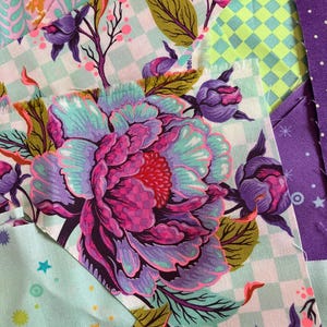 Puede incluir: Primer plano de piezas de tela coloridas con estampados florales y geométricos. La pieza dominante presenta una gran peonía en tonos morado, rosa y turquesa. Otras telas incluyen diseños a cuadros, lunares y estampados en varios colores.