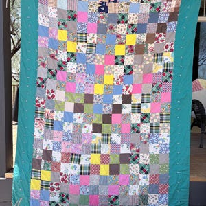 Può includere: Una trapunta patchwork composta da una griglia di quadrati di tessuto multicolore con vari motivi, tra cui fiori, quadri e colori a tinta unita. La trapunta è incorniciata da un ampio bordo verde acqua ed è appesa con mollette di legno.