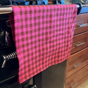 May include: A pink and brown checkered kitchen towel hanging on a stove. The towel has a classic gingham pattern, with alternating squares of bright pink and a darker brown. The towel is made of a soft, woven fabric.