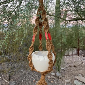 Puede incluir: Un colgador de plantas de macramé hecho a mano con una maceta de cerámica blanca. El diseño de cuerda marrón presenta detalles rojos y una borla en la parte inferior. El colgador de plantas está suspendido de una rama de árbol, mostrando un estilo bohemio.