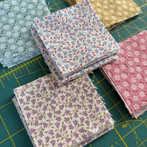 Puede incluir: Varias pilas de pequeños trozos de tela cuadrados con estampados florales en varios colores. Las telas incluyen azul claro, crema, rosa y dorado, cada una con un diseño floral diferente. Los cuadrados están apilados ordenadamente sobre una esterilla de corte verde.