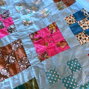 May include: A patchwork quilt featuring a light blue fabric grid separating various square blocks. The blocks include patterns such as pink floral, brown paisley, green plaid, blue polka dots, and multi-colored floral prints in a variety of colors.