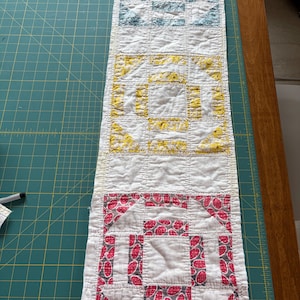 May include: A rectangular, handmade quilt runner with three distinct sections. Each section features a square-in-a-square design in blue, yellow, and red patterns on a white background. The runner is likely made of cotton and shows signs of wear.