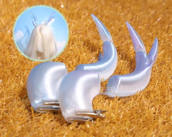 May include: Iridescent blue and white hair clips with a horn-like design. The clips are arranged on a bed of golden-brown artificial grass. A circular inset shows a person wearing a wig with similar horn-like hair accessories.
